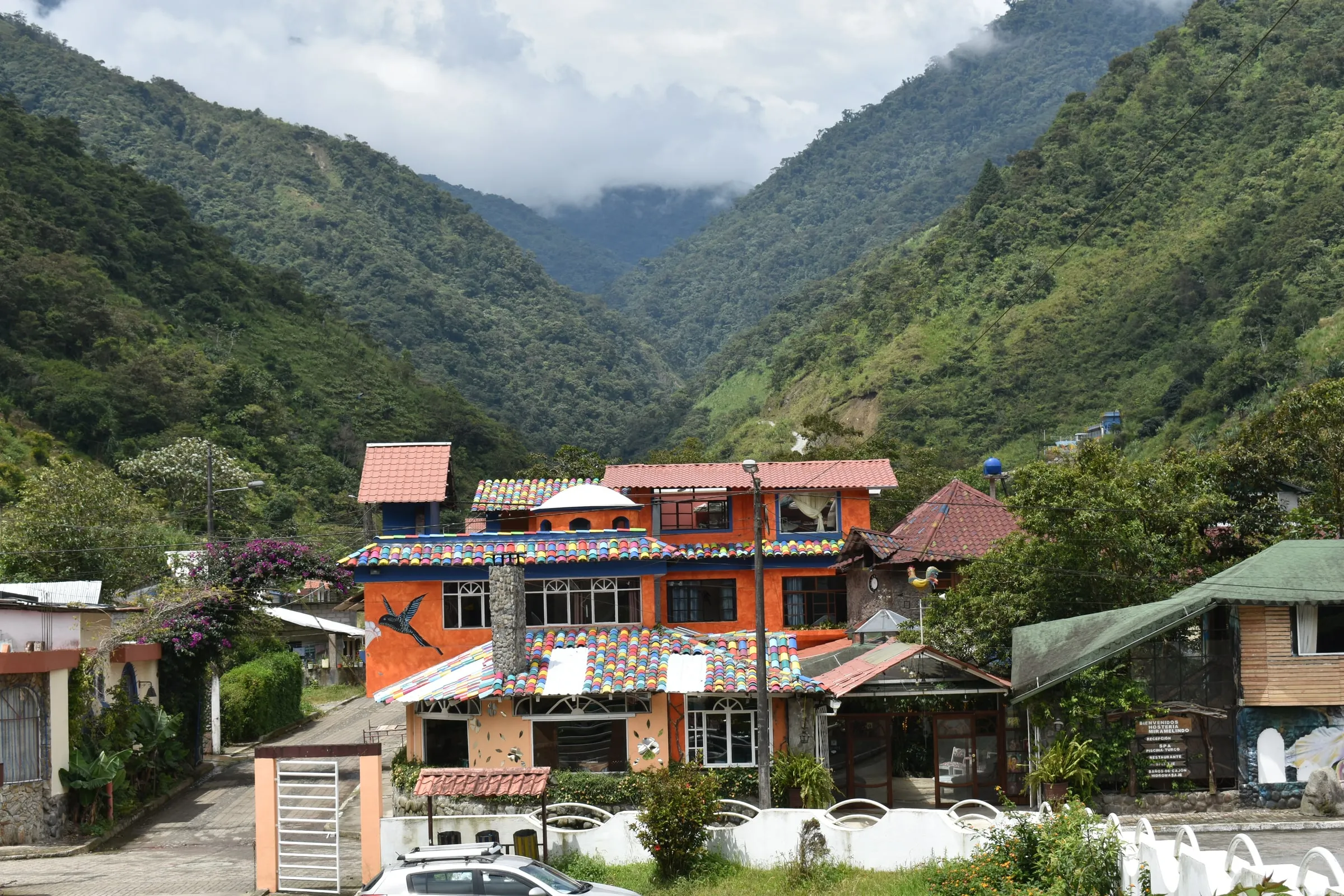 Fachada colorida de Hotel Spa Miramelindo, con techos de mosaico y un colibrí pintado, rodeada de montañas cubiertas de bosque en Río Verde, Baños de Agua Santa